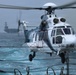 A white civilian helicopter registered N597AC, marked 'Rodimus Prime' with number '04', is hovering or landing on a naval vessel deck over choppy open ocean. In the background, a large aircraft carrier is visible through hazy conditions. The scene appears to depict a helicopter transfer or resupply operation between vessels at sea.