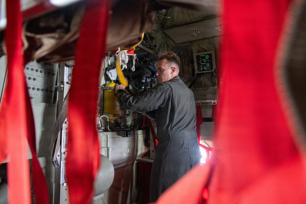 A person in a gray flight suit is working inside what appears to be a military or rescue aircraft, handling yellow and black equipment mounted on the interior wall. The image is shot through red safety straps or harnesses in the foreground, which partially obscure the view. Various avionics displays, panels, and medical/rescue equipment are visible on the aircraft interior walls.