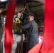 A person in a gray flight suit is working inside what appears to be a military or rescue aircraft, handling yellow and black equipment mounted on the interior wall. The image is shot through red safety straps or harnesses in the foreground, which partially obscure the view. Various avionics displays, panels, and medical/rescue equipment are visible on the aircraft interior walls.