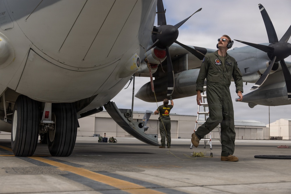 Military personnel are conducting pre-flight or post-flight inspection and maintenance on a large turboprop aircraft on a tarmac. A flight crew member in an olive green flight suit walks beneath the aircraft while a ground crew member in a military t-shirt works near the fuselage with a ladder and rope/cable. The setting appears to be a military air base with hangars visible in the background.