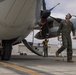 Military personnel are conducting pre-flight or post-flight inspection and maintenance on a large turboprop aircraft on a tarmac. A flight crew member in an olive green flight suit walks beneath the aircraft while a ground crew member in a military t-shirt works near the fuselage with a ladder and rope/cable. The setting appears to be a military air base with hangars visible in the background.
