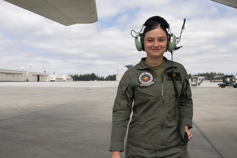 A female military aviator or aircrew member stands on a flight line apron wearing an olive green flight suit and aviation headset. She is smiling at the camera with aircraft infrastructure and hangars visible in the background. The setting appears to be a military air station or airfield.