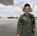 A female military aviator or aircrew member stands on a flight line apron wearing an olive green flight suit and aviation headset. She is smiling at the camera with aircraft infrastructure and hangars visible in the background. The setting appears to be a military air station or airfield.