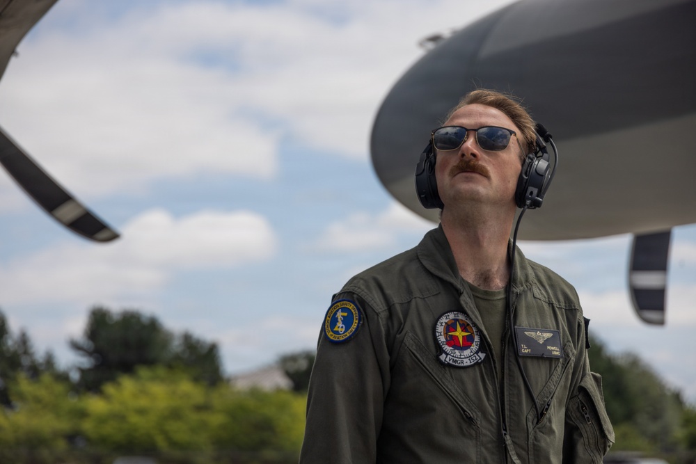 A military aviator in an olive green flight suit stands near a large aircraft, looking upward. He is wearing aviator sunglasses, a communications headset, and displays multiple patches on his uniform including a name tag reading 'POWELL' with rank 'CAPT' and branch 'USMC'. The image is taken from a low angle with the aircraft's fuselage or nacelle visible in the background against a partly cloudy sky.