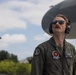 A military aviator in an olive green flight suit stands near a large aircraft, looking upward. He is wearing aviator sunglasses, a communications headset, and displays multiple patches on his uniform including a name tag reading 'POWELL' with rank 'CAPT' and branch 'USMC'. The image is taken from a low angle with the aircraft's fuselage or nacelle visible in the background against a partly cloudy sky.