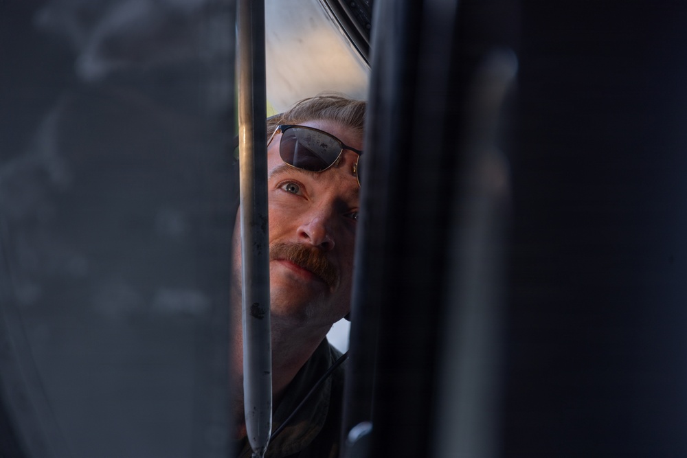 A man with a mustache and sunglasses pushed up on his forehead peers through a narrow gap between what appear to be vehicle or machinery panels. The shot is taken from a tight, constrained angle, framing his face between dark metallic surfaces. The lighting highlights his face while the surrounding elements remain in shadow.