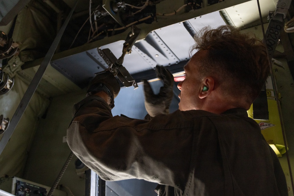 A technician or military maintainer is working on the interior mechanical components of what appears to be a large military aircraft, likely a cargo or transport plane. The individual is reaching upward to manipulate hardware, cables, or fasteners in the overhead structural area. The image is taken from a low angle inside the aircraft, emphasizing the complexity of the internal framework.