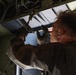A technician or military maintainer is working on the interior mechanical components of what appears to be a large military aircraft, likely a cargo or transport plane. The individual is reaching upward to manipulate hardware, cables, or fasteners in the overhead structural area. The image is taken from a low angle inside the aircraft, emphasizing the complexity of the internal framework.