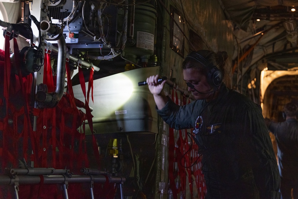 A military aircrew member wearing a flight suit, headset, and safety glasses is conducting an inspection inside what appears to be a large military transport aircraft, likely a C-130 or similar airlifter. The individual is using a flashlight to illuminate equipment mounted on the interior wall of the aircraft. Red cargo netting and various mechanical components are visible throughout the cargo bay.