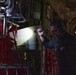 A military aircrew member wearing a flight suit, headset, and safety glasses is conducting an inspection inside what appears to be a large military transport aircraft, likely a C-130 or similar airlifter. The individual is using a flashlight to illuminate equipment mounted on the interior wall of the aircraft. Red cargo netting and various mechanical components are visible throughout the cargo bay.