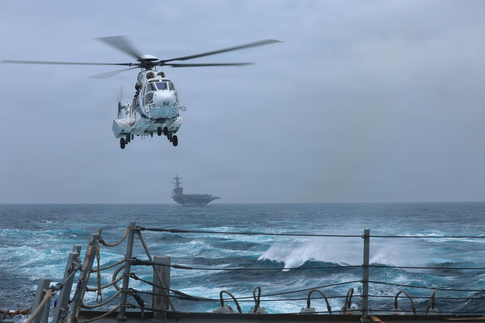 A military helicopter numbered '04' hovers over the deck of a naval vessel in rough open-ocean conditions. In the background, a large aircraft carrier is visible through hazy, overcast skies. The scene depicts an active naval operation with choppy seas and low visibility weather.