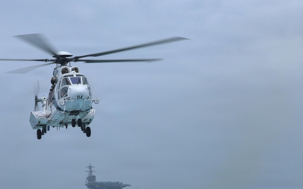 A military helicopter numbered '04' hovers over the deck of a naval vessel in rough open-ocean conditions. In the background, a large aircraft carrier is visible through hazy, overcast skies. The scene depicts an active naval operation with choppy seas and low visibility weather.