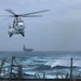 A military helicopter numbered '04' hovers over the deck of a naval vessel in rough open-ocean conditions. In the background, a large aircraft carrier is visible through hazy, overcast skies. The scene depicts an active naval operation with choppy seas and low visibility weather.