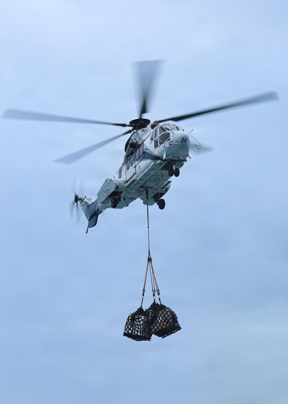 A military helicopter, marked with the number '04' and appearing light gray/white in color, is airborne against an overcast blue-gray sky. It is conducting a vertical replenishment (VERTREP) operation, carrying two cargo net loads suspended beneath the fuselage via a sling line. The rotor blades show motion blur, indicating active flight.