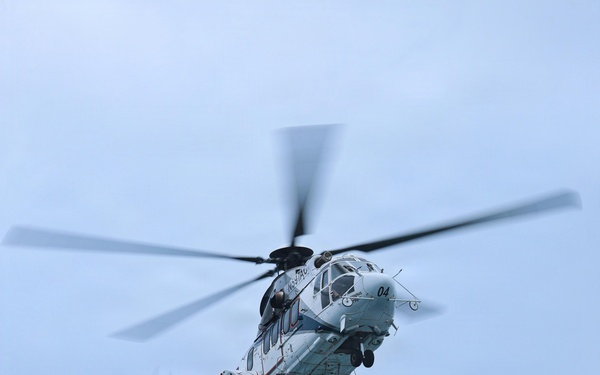 A military helicopter, marked with the number '04' and appearing light gray/white in color, is airborne against an overcast blue-gray sky. It is conducting a vertical replenishment (VERTREP) operation, carrying two cargo net loads suspended beneath the fuselage via a sling line. The rotor blades show motion blur, indicating active flight.