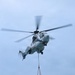 A military helicopter, marked with the number '04' and appearing light gray/white in color, is airborne against an overcast blue-gray sky. It is conducting a vertical replenishment (VERTREP) operation, carrying two cargo net loads suspended beneath the fuselage via a sling line. The rotor blades show motion blur, indicating active flight.