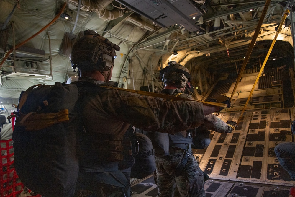 Military personnel equipped with tactical gear, helmets with night-vision mounts, and parachute rigs are positioned at the open rear ramp of a large military cargo aircraft, likely a C-130 or C-17. They appear to be preparing for or in the final moments before a parachute jump. A yellow static line or safety strap is visible across the frame, consistent with standard military airborne operations.