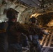 Military personnel equipped with tactical gear, helmets with night-vision mounts, and parachute rigs are positioned at the open rear ramp of a large military cargo aircraft, likely a C-130 or C-17. They appear to be preparing for or in the final moments before a parachute jump. A yellow static line or safety strap is visible across the frame, consistent with standard military airborne operations.