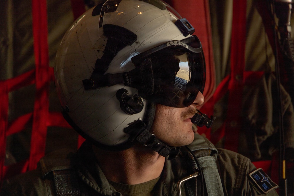 A military aviator or aircrew member is photographed in profile wearing a flight helmet with a tinted visor. The visor reflects what appears to be an aerial landscape, suggesting the aircraft is airborne. The subject is dressed in an olive drab flight suit with a visible patch on the shoulder.
