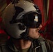 A military aviator or aircrew member is photographed in profile wearing a flight helmet with a tinted visor. The visor reflects what appears to be an aerial landscape, suggesting the aircraft is airborne. The subject is dressed in an olive drab flight suit with a visible patch on the shoulder.