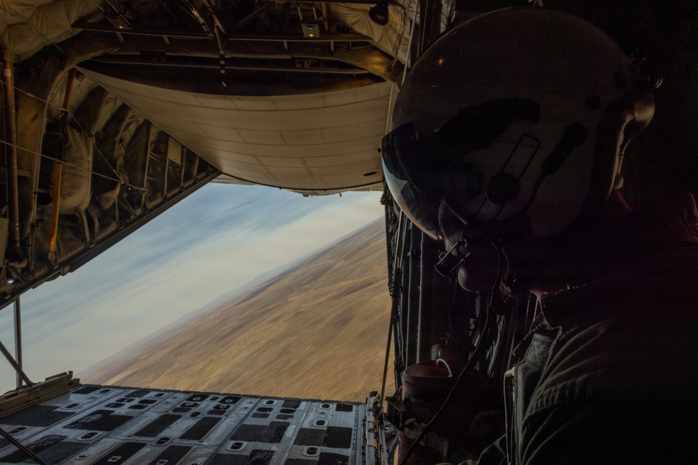 A military aircrew member wearing a flight helmet and communications headset is positioned at the open rear cargo ramp of a military transport aircraft, likely a C-130 Hercules or similar platform. The aircraft is flying at low altitude over an arid, desert terrain visible through the open ramp. The image is taken from inside the aircraft looking outward, capturing both the crew member and the expansive landscape below.