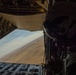 A military aircrew member wearing a flight helmet and communications headset is positioned at the open rear cargo ramp of a military transport aircraft, likely a C-130 Hercules or similar platform. The aircraft is flying at low altitude over an arid, desert terrain visible through the open ramp. The image is taken from inside the aircraft looking outward, capturing both the crew member and the expansive landscape below.