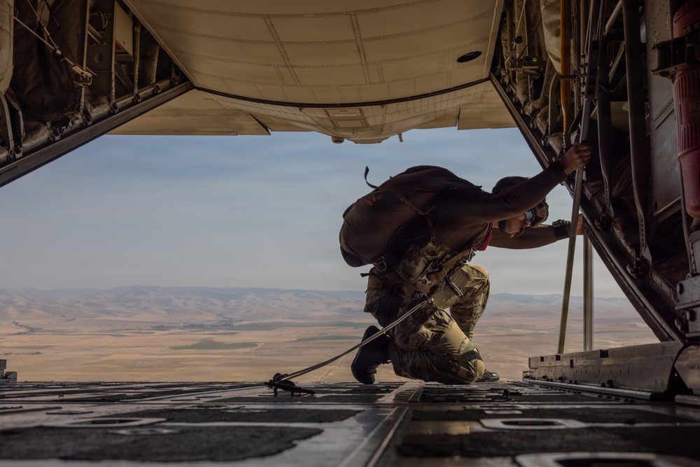A military personnel wearing full combat gear and a parachute pack is kneeling on the open rear ramp of a military cargo aircraft, likely a C-130 Hercules, in flight. The individual appears to be inspecting or adjusting equipment near the edge of the ramp. Below and behind the aircraft, an arid, semi-desert landscape with rolling terrain is visible.