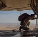 A military personnel wearing full combat gear and a parachute pack is kneeling on the open rear ramp of a military cargo aircraft, likely a C-130 Hercules, in flight. The individual appears to be inspecting or adjusting equipment near the edge of the ramp. Below and behind the aircraft, an arid, semi-desert landscape with rolling terrain is visible.