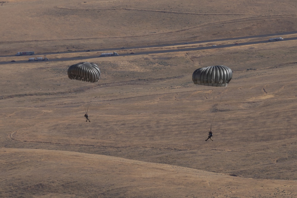Two military paratroopers are descending toward arid, rolling terrain using dark olive/gray round parachutes. The scene appears to be a military airborne training or operational jump over a dry, open landscape. A road with vehicles is visible in the background, suggesting proximity to a base or training area.
