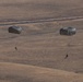 Two military paratroopers are descending toward arid, rolling terrain using dark olive/gray round parachutes. The scene appears to be a military airborne training or operational jump over a dry, open landscape. A road with vehicles is visible in the background, suggesting proximity to a base or training area.