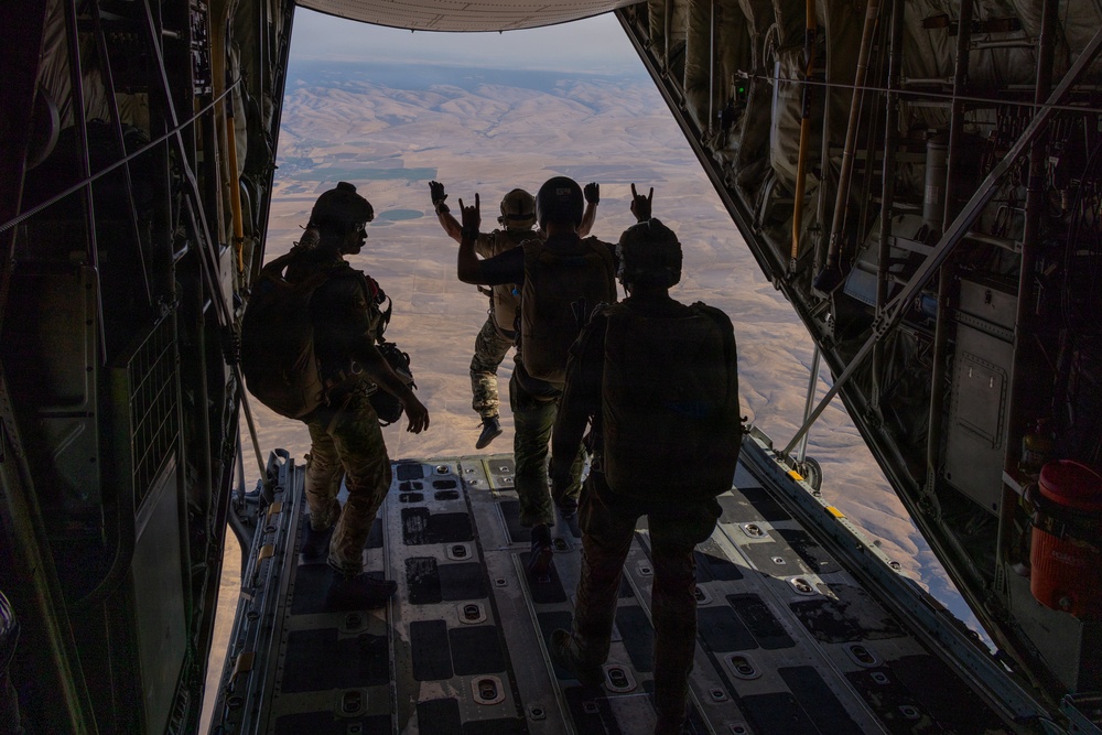 Four military personnel in tactical gear are standing at the open rear ramp of a large military cargo aircraft, likely a C-130 Hercules, at altitude. Two individuals in the center are making hand gestures (rock/horns signs) toward the open ramp with an arid, mountainous landscape visible far below. The image is taken from inside the aircraft looking outward, creating a dramatic silhouette effect.