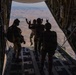 Four military personnel in tactical gear are standing at the open rear ramp of a large military cargo aircraft, likely a C-130 Hercules, at altitude. Two individuals in the center are making hand gestures (rock/horns signs) toward the open ramp with an arid, mountainous landscape visible far below. The image is taken from inside the aircraft looking outward, creating a dramatic silhouette effect.