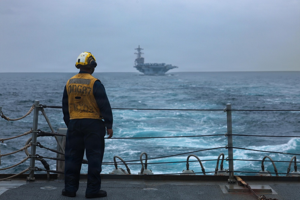 A U.S. Navy sailor stands on the deck of a destroyer or guided-missile destroyer, wearing a yellow safety vest marked 'DDG 87' and a white helmet with hearing protection, observing a large aircraft carrier in the distance. The scene appears to be taken during overcast, rough sea conditions. This appears to be a naval formation or escort operation at sea.
