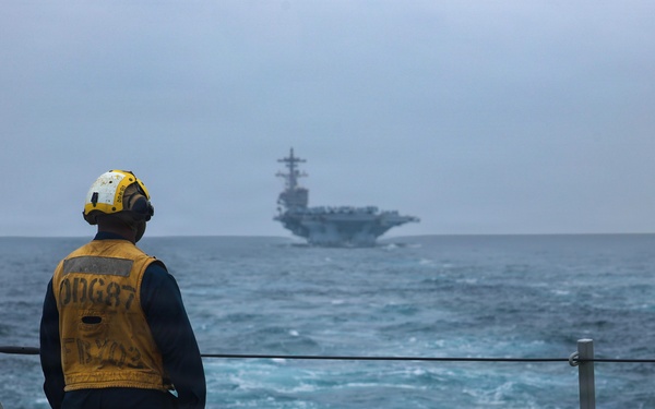 A U.S. Navy sailor stands on the deck of a destroyer or guided-missile destroyer, wearing a yellow safety vest marked 'DDG 87' and a white helmet with hearing protection, observing a large aircraft carrier in the distance. The scene appears to be taken during overcast, rough sea conditions. This appears to be a naval formation or escort operation at sea.