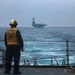 A U.S. Navy sailor stands on the deck of a destroyer or guided-missile destroyer, wearing a yellow safety vest marked 'DDG 87' and a white helmet with hearing protection, observing a large aircraft carrier in the distance. The scene appears to be taken during overcast, rough sea conditions. This appears to be a naval formation or escort operation at sea.