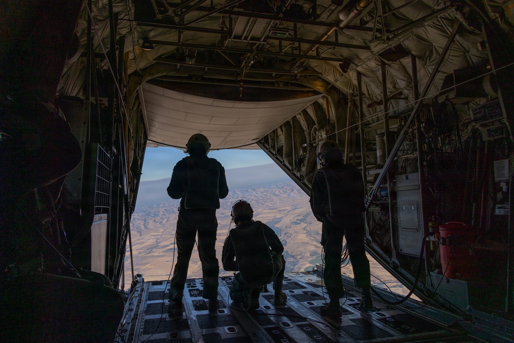 Three military personnel are silhouetted against an open cargo ramp of a large military transport aircraft, looking out over an arid, mountainous landscape below. The individuals are wearing flight gear and communications headsets, suggesting they are aircrew or loadmasters. The scene appears to be taken during flight at significant altitude over a desert or semi-arid terrain.