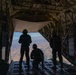 Three military personnel are silhouetted against an open cargo ramp of a large military transport aircraft, looking out over an arid, mountainous landscape below. The individuals are wearing flight gear and communications headsets, suggesting they are aircrew or loadmasters. The scene appears to be taken during flight at significant altitude over a desert or semi-arid terrain.