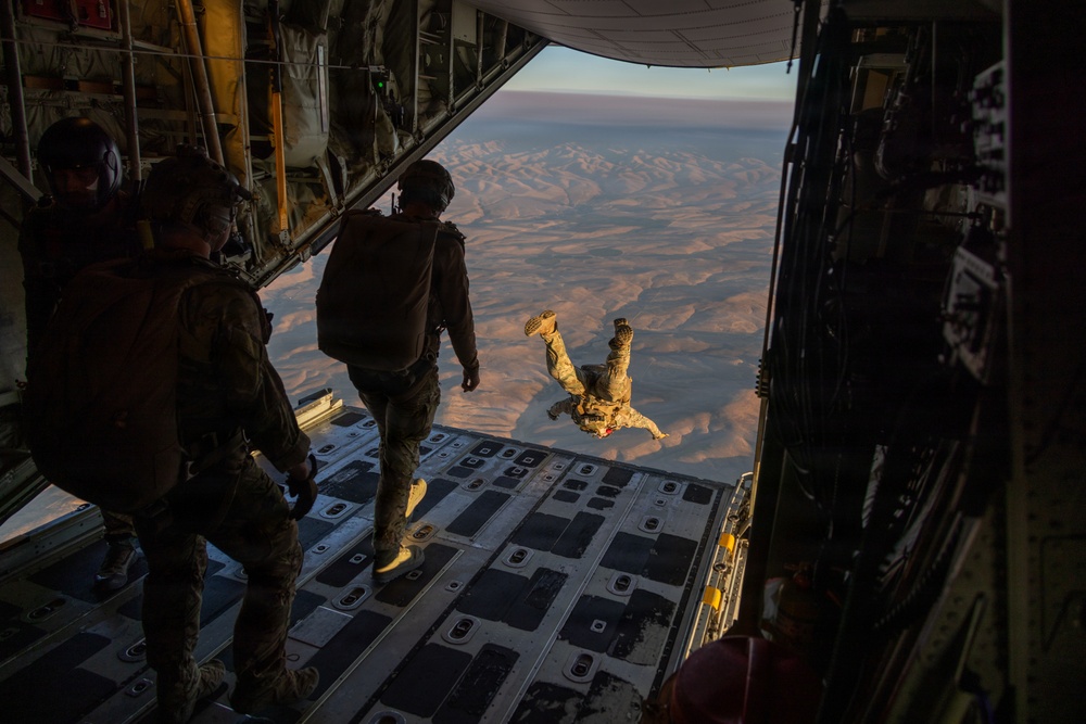 Military personnel are conducting a parachute jump operation from the open rear ramp of a large military cargo aircraft, likely a C-130 or C-17, over an arid desert landscape. One jumper has already exited the aircraft and is visible in freefall in a head-down position, while two other personnel stand on the ramp observing. The scene is illuminated by warm golden-hour light, suggesting either dawn or dusk conditions.