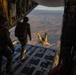 Military personnel are conducting a parachute jump operation from the open rear ramp of a large military cargo aircraft, likely a C-130 or C-17, over an arid desert landscape. One jumper has already exited the aircraft and is visible in freefall in a head-down position, while two other personnel stand on the ramp observing. The scene is illuminated by warm golden-hour light, suggesting either dawn or dusk conditions.