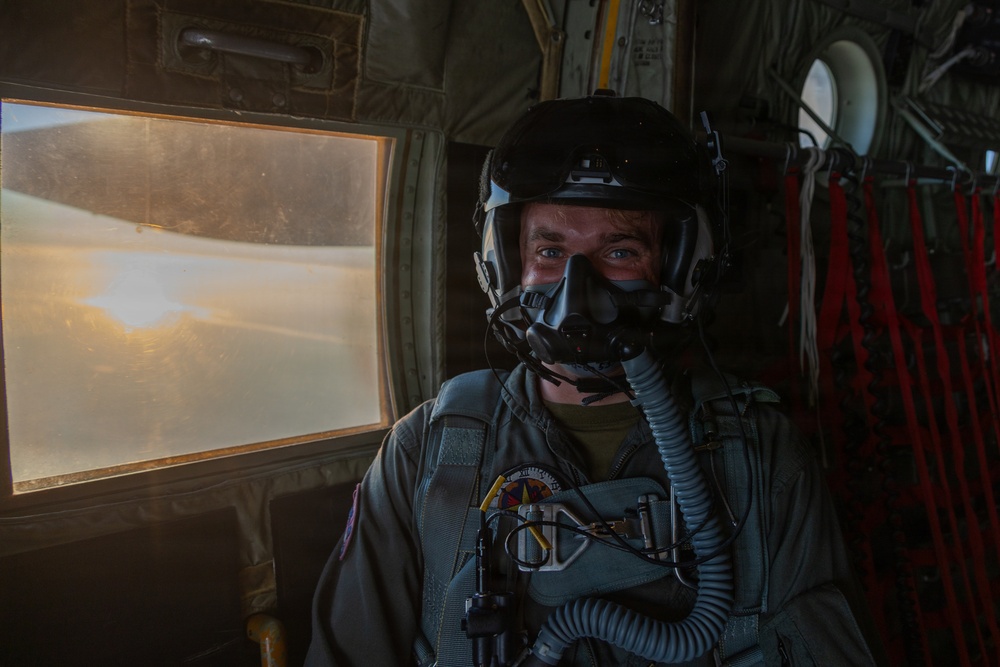A military aviator is seated inside a large aircraft, wearing full flight gear including a helmet with night vision goggle mount and an oxygen mask. The interior of the aircraft shows red cargo netting and structural elements typical of a military transport or tanker aircraft. Warm golden light from a window illuminates the scene, suggesting flight at sunset or sunrise.