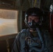 A military aviator is seated inside a large aircraft, wearing full flight gear including a helmet with night vision goggle mount and an oxygen mask. The interior of the aircraft shows red cargo netting and structural elements typical of a military transport or tanker aircraft. Warm golden light from a window illuminates the scene, suggesting flight at sunset or sunrise.