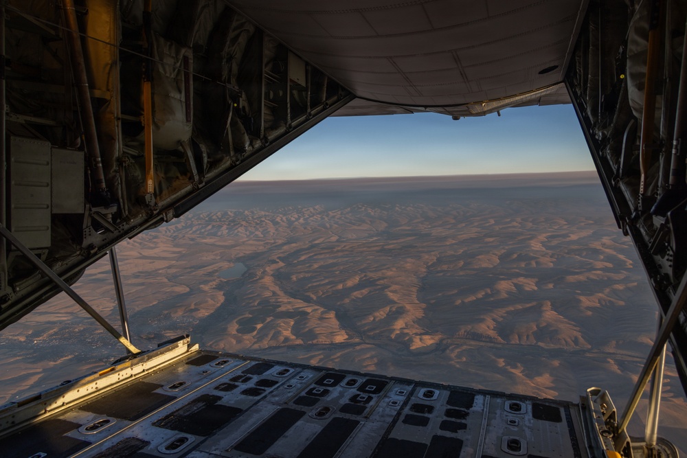 The image is taken from inside a military cargo aircraft, looking out through an open rear cargo ramp door. Below, an arid desert landscape with rolling hills and dry riverbeds is visible, illuminated by low-angle golden light suggesting either sunrise or sunset. The aircraft interior shows structural framing, hydraulic lines, and the partially lowered cargo ramp.
