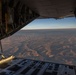 The image is taken from inside a military cargo aircraft, looking out through an open rear cargo ramp door. Below, an arid desert landscape with rolling hills and dry riverbeds is visible, illuminated by low-angle golden light suggesting either sunrise or sunset. The aircraft interior shows structural framing, hydraulic lines, and the partially lowered cargo ramp.