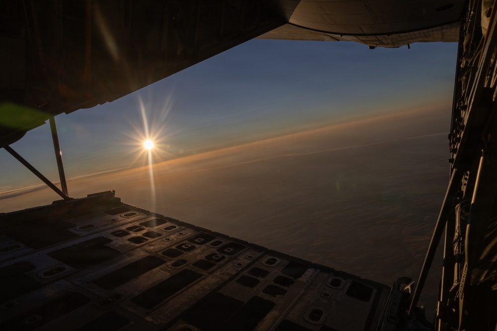 The image is taken from inside a military cargo aircraft, looking out through an open rear cargo ramp door at a sunrise or sunset on the horizon. The aircraft is flying at altitude above a cloud layer, with warm orange and golden light emanating from the sun near the horizon. The interior of the cargo bay floor with its tie-down track system is visible in the foreground.