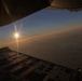 The image is taken from inside a military cargo aircraft, looking out through an open rear cargo ramp door at a sunrise or sunset on the horizon. The aircraft is flying at altitude above a cloud layer, with warm orange and golden light emanating from the sun near the horizon. The interior of the cargo bay floor with its tie-down track system is visible in the foreground.