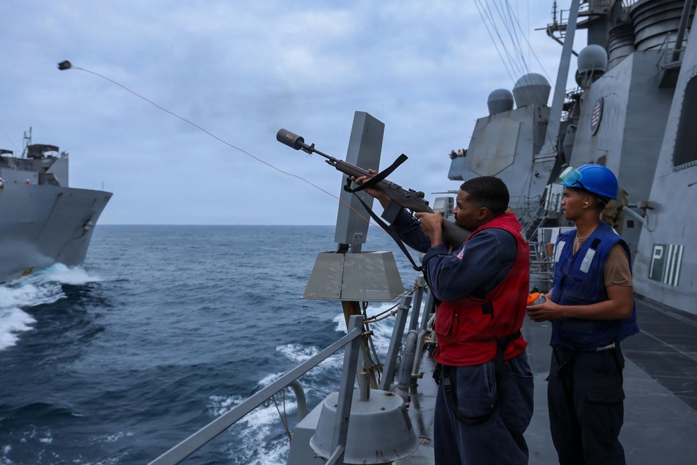 Two naval personnel are conducting a replenishment-at-sea (RAS) operation aboard a U.S. Navy warship. One sailor in a red vest is firing a line-throwing gun to establish a connection between two vessels sailing in close proximity. A projectile and red messenger line are visibly in flight toward the adjacent ship.