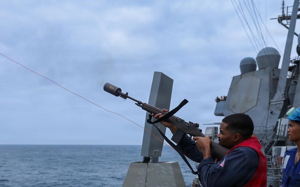 Two naval personnel are conducting a replenishment-at-sea (RAS) operation aboard a U.S. Navy warship. One sailor in a red vest is firing a line-throwing gun to establish a connection between two vessels sailing in close proximity. A projectile and red messenger line are visibly in flight toward the adjacent ship.
