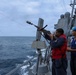 Two naval personnel are conducting a replenishment-at-sea (RAS) operation aboard a U.S. Navy warship. One sailor in a red vest is firing a line-throwing gun to establish a connection between two vessels sailing in close proximity. A projectile and red messenger line are visibly in flight toward the adjacent ship.