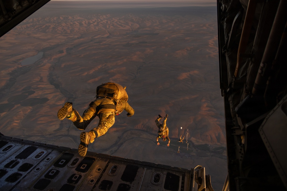 Military personnel are conducting a high-altitude parachute jump from the open rear ramp of a large military transport aircraft, likely a C-130 or C-17. The jumpers are wearing full tactical gear and parachute rigs in tan/desert camouflage coloring. The terrain below appears to be an arid desert landscape, photographed during golden hour lighting conditions.