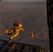Military personnel are conducting a high-altitude parachute jump from the open rear ramp of a large military transport aircraft, likely a C-130 or C-17. The jumpers are wearing full tactical gear and parachute rigs in tan/desert camouflage coloring. The terrain below appears to be an arid desert landscape, photographed during golden hour lighting conditions.